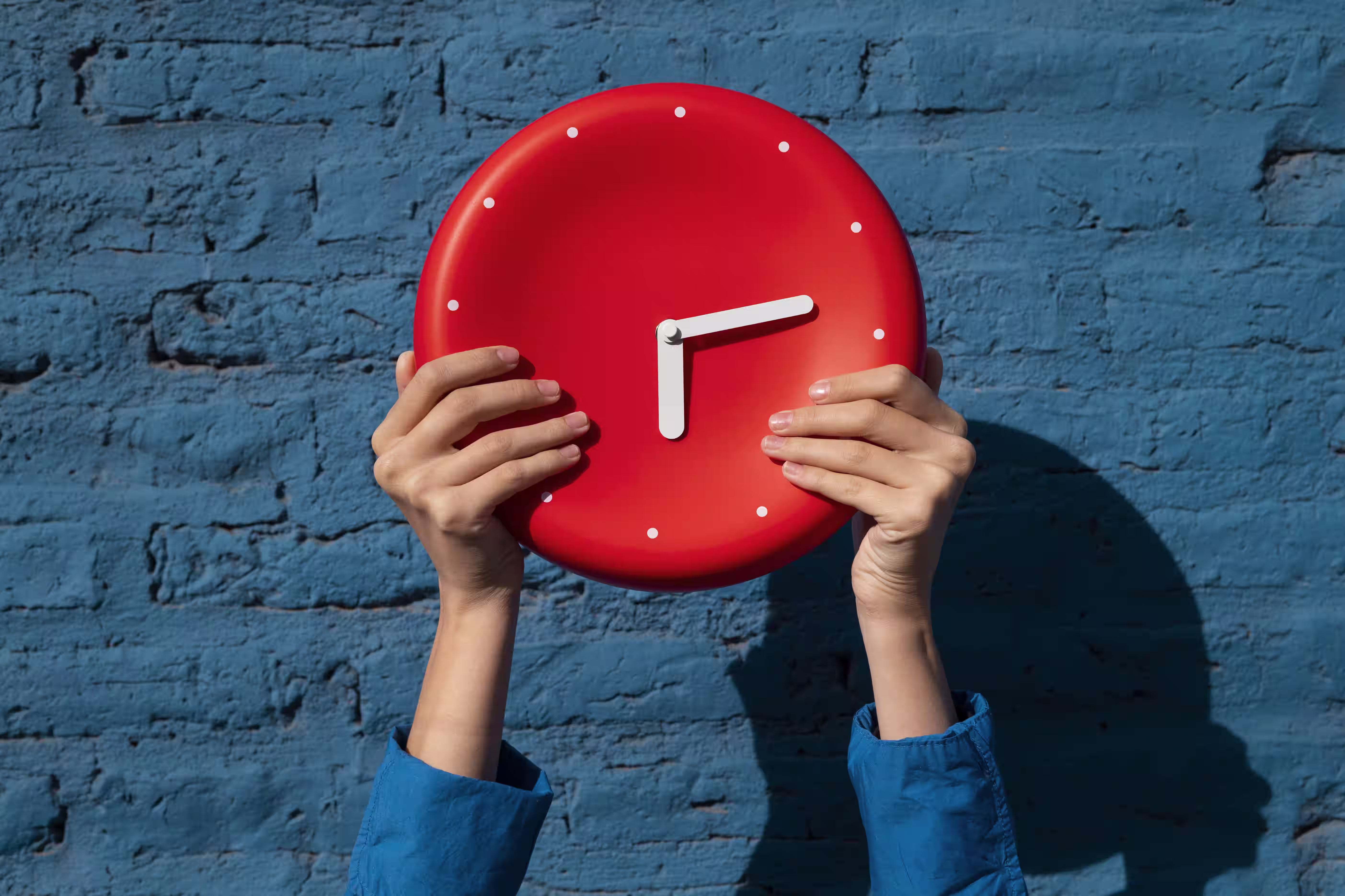 Hands holding a bright red analog clock against a blue brick wall background