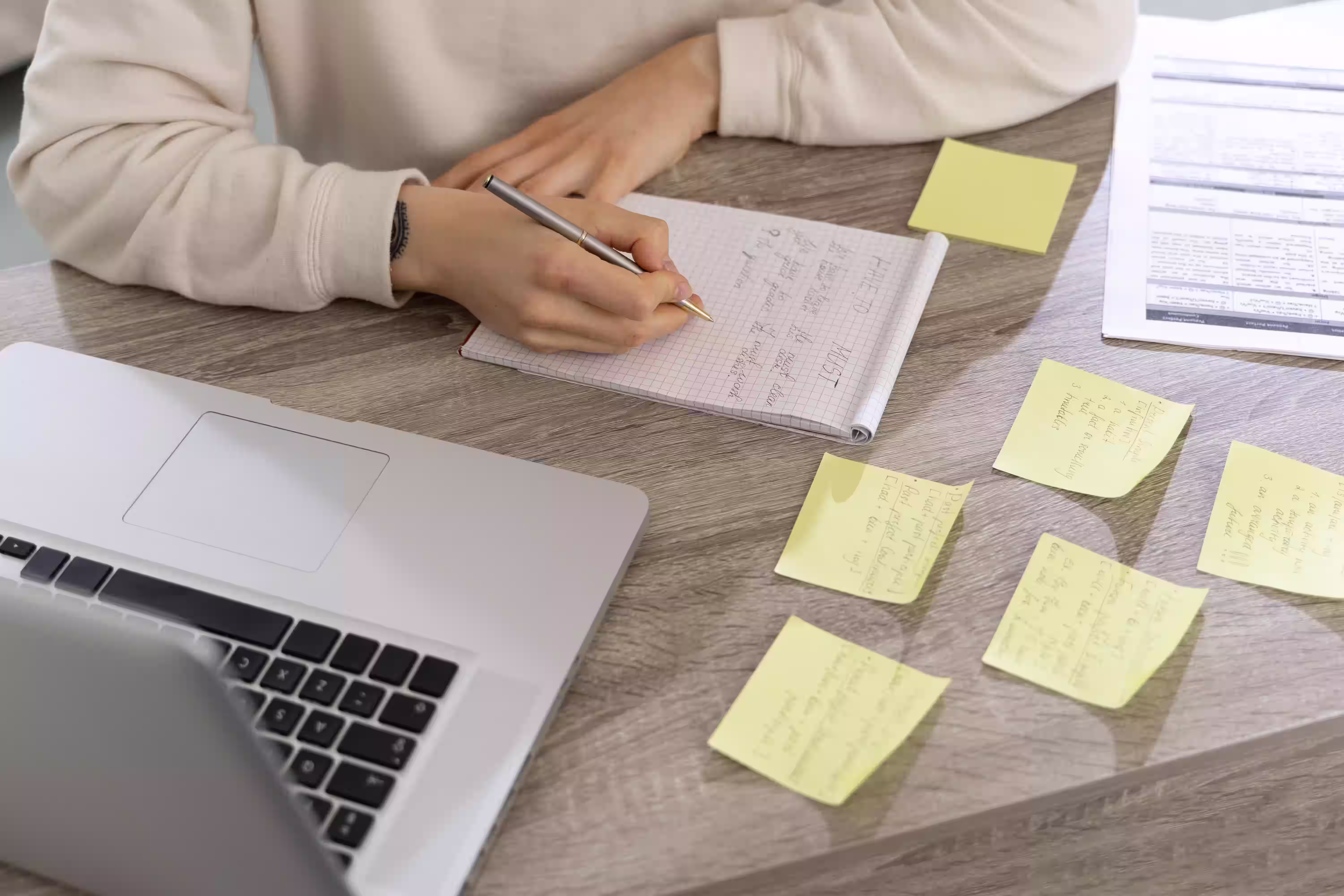 Person writing notes in grid notebook with sticky notes and documents on wooden desk