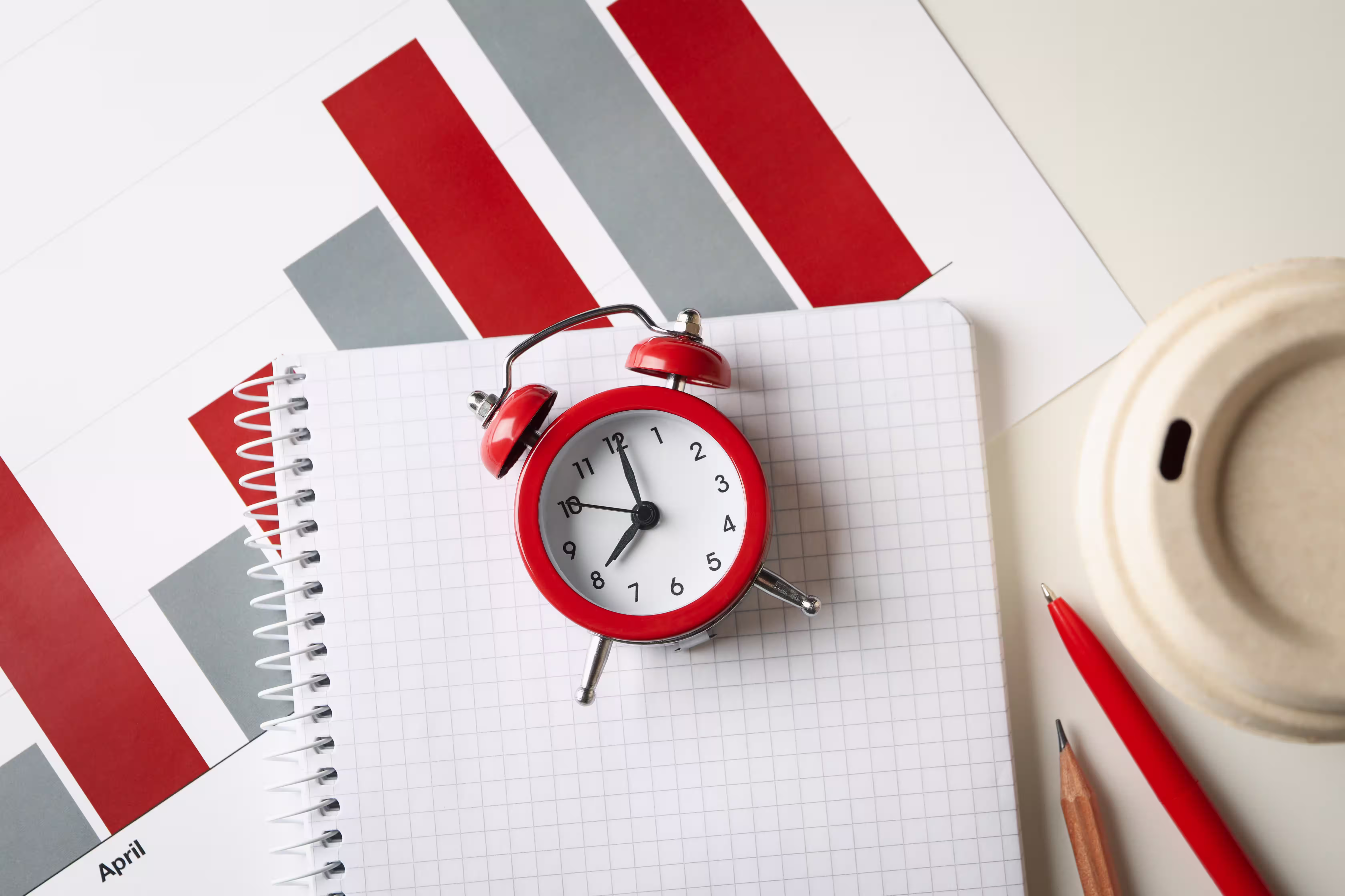 Red alarm clock on a gridded notebook with planning documents and stationery items