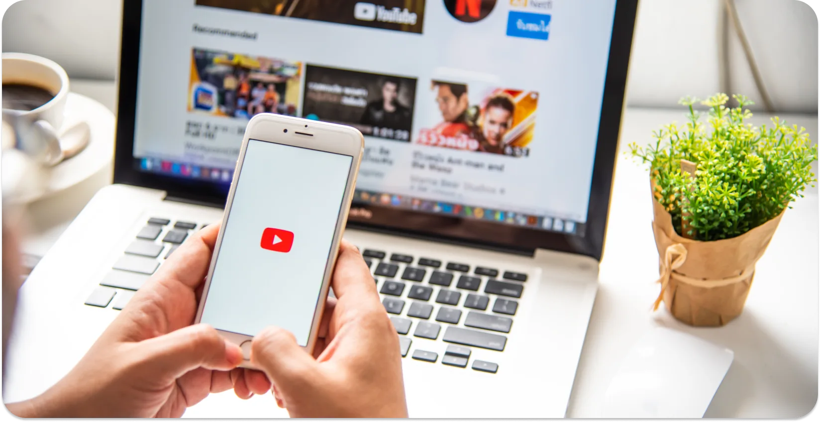 Osoba koja drži pametni telefon s prikazanim logotipom YouTube, s prijenosnim računalom otvorenim za YouTube u pozadini.